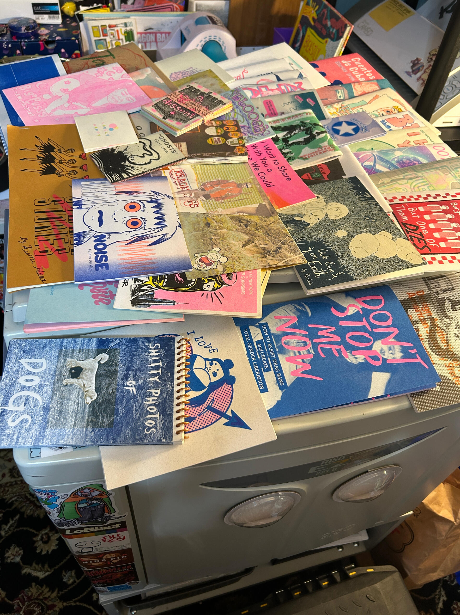 A photograph of many zines printed with a Risograph, layered on the top of our EZ 221 U, which has big googly eyes on it.